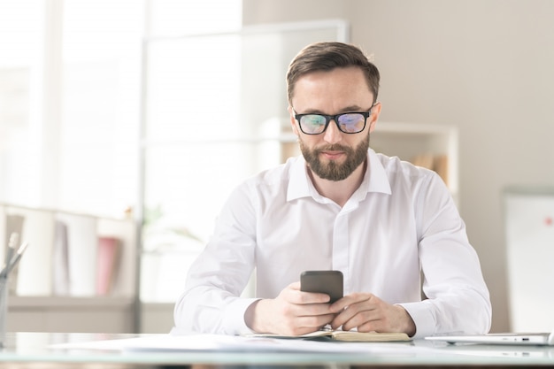 Серьезный молодой офис-менеджер или предприниматель в белой рубашке сидит на рабочем месте и прокручивает в своем смартфоне