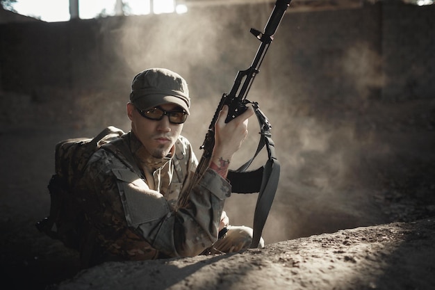 A serious young soldier of an independent state in camouflage uniform with a large backpack on his back in tactical glasses and in a cap reloads a machine gun while in cover