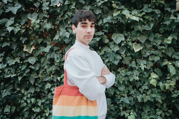 Giovane uomo spagnolo serio che guarda l'obbiettivo con una borsa tote lgtbi che guarda l'obbiettivo sorridente per strada, indossando un maglione bianco. movimento dell'orgoglio gay lgbti e stile moderno. concetto gay