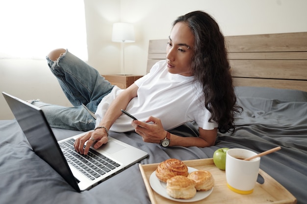 Serious young Vietnamese man with wavy hair lying on bed with tray with buns, coffee and apple and using modern devices in bedroom