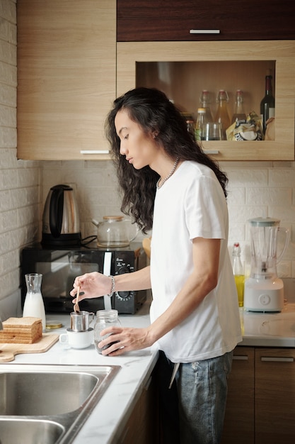 serious young vietnamese man with wavy hair standing at kitchen counter and making coffee in morning