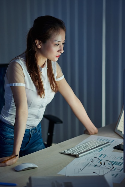 Serious young Vietnamese project manager looking at glowing screen of computer in dark office