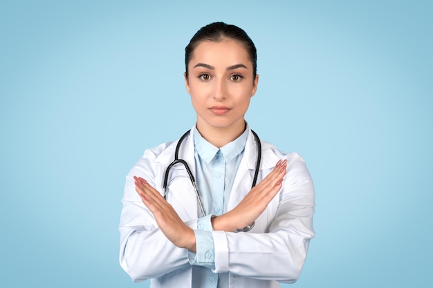 Serious young woman doctor in white coat making x sign with her arms signaling denial or prohibition