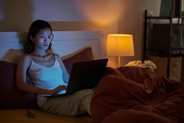 写真 パジャマを着てベッドに座り、夜遅くまでラップトップで作業する深刻な若い女性