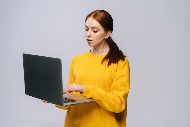 ラップトップコンピューターを保持し、孤立した灰色の背景に入力する深刻な若い女性の学生スタジオのコピースペースで顔の表情を感情的に示すかなり赤毛の女性モデル