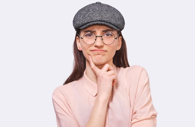 Serious young woman wearing casual outfit and round transparent spectacles frowning her face and looking to the camera poses against white studio background People emotions concept