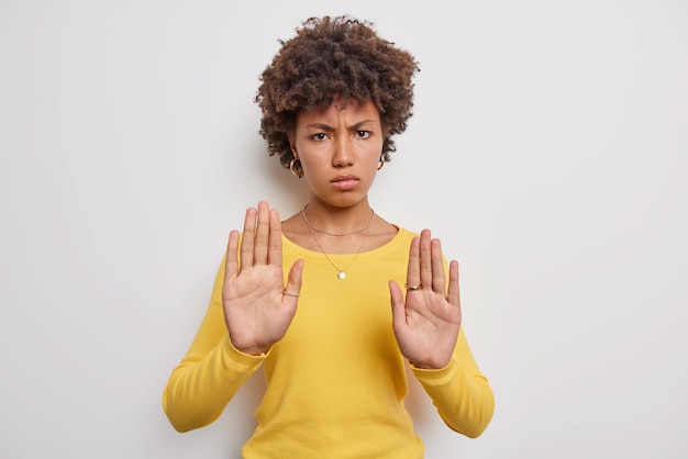 Serious young woman with curly hair keeps palms raised towards camera has angry expression demands not to come closer wears yellow casual jumper isolated over white background makes protest