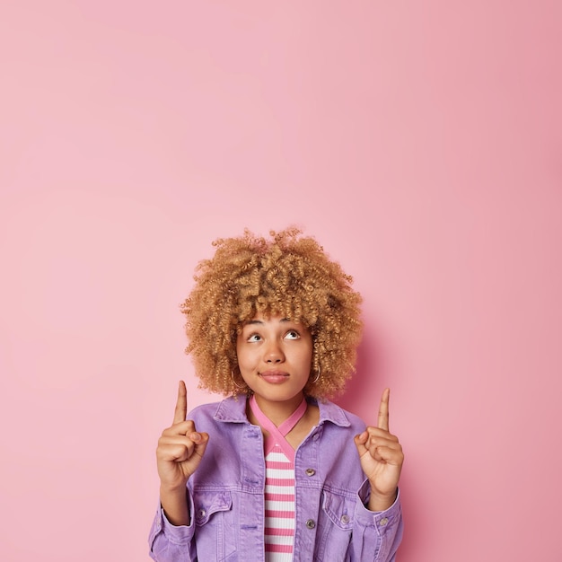 Serious young woman with curly hair points overhead with both\
index fingers advertises product dressed in purple jacket shows\
promo isolated over pink background look at this logo or\
banner