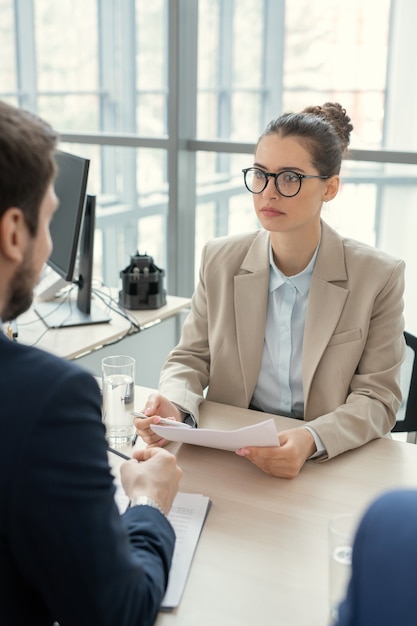 Serious young woman with hair bun sitting at table and analyzing contract with lawyer in office