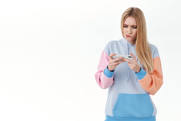 Серьезно выглядящая привлекательная блондинка загружает новую гоночную игру, держа мобильный телефон горизонтально, дуется и хмурится, пытаясь пройти сложный уровень в онлайн-аркаде на белом фоне
