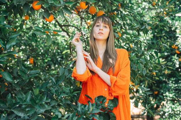 Seriously beautiful girl in orange dress is posing to camera by holding up and together hands in orange garden