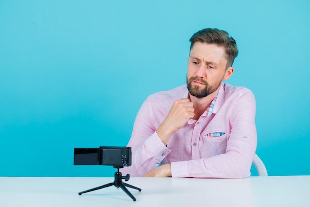 Seriously blogger man is posing to his mini camera by holding collar of shirt on blue background