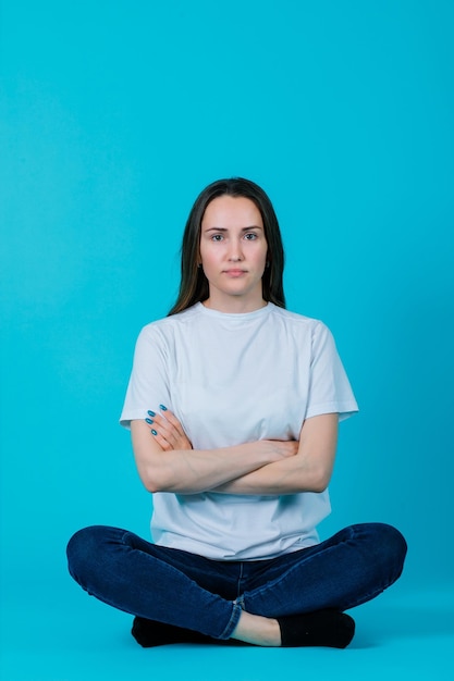 Seriously girl is crossing arms by sitting on floor on blue background