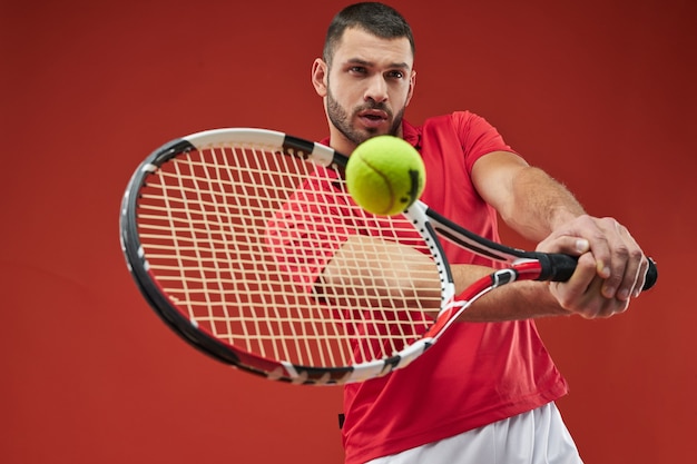 赤い背景で隔離のテニスをしている赤いシャツの真剣に強いアスリート男性
