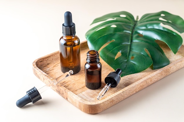 Serum or essential oil in brown glass dropper bottles on wooden tray with monstera leaf, beige background. Natural Organic Spa Cosmetic concept. Front view.