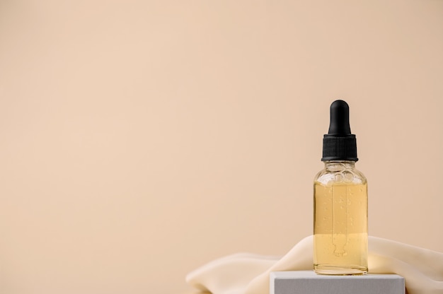 Serum in a glass bottle with a pipette on a gray stand with a silk cloth. Essential oil for the care of female skin. Skin care, moisturizing beauty product on a beige background. Copy space.