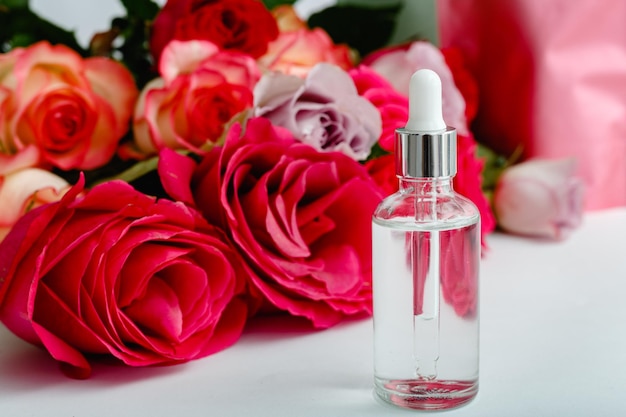 Serum on white table floral background. Flower red pink roses natural beauty product. Skin care,body treatment. Glass cosmetic bottle with rose copy space.