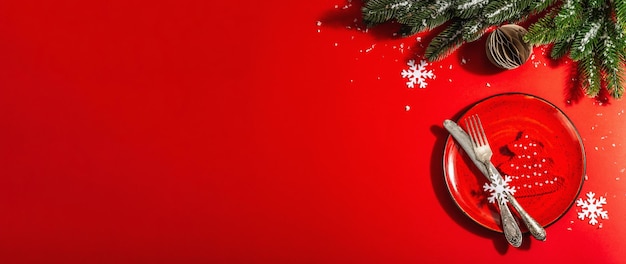 Served Christmas table setting in red tones with vintage cutlery. Evergreen branches, crocheted fir trees, artificial snow. Modern hard light, dark shadow. Flat lay, red background, top view