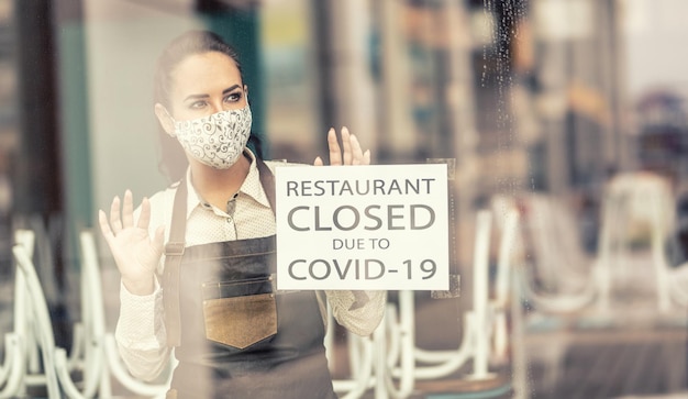 Serveerster in een gesloten restaurant vanwege Covid19 draagt gezichtsmasker en kijkt uit het raam