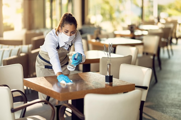 Serveerster met een gezichtsmasker en handschoenen die tafels schoonmaakt met ontsmettingsmiddel in een café