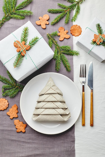 Servet in de vorm van een kerstboom op een bord op wit tafelkleed met geschenken en versieringen met spartakjes en peperkoekkoekjes