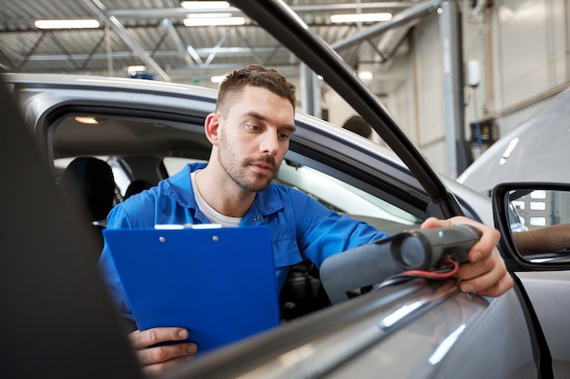 service, repair, maintenance and people concept - mechanic man with automotive diagnostic scanner and clipboard checking car system at workshop