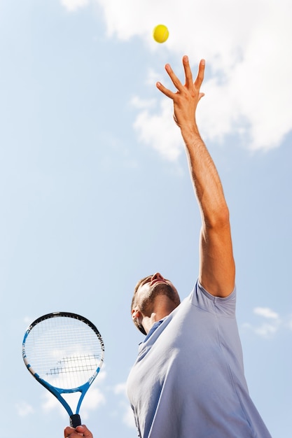 공을 제공합니다. 푸른 하늘에 서 있는 동안 공을 제공하는 테니스 선수의 낮은 각도 보기