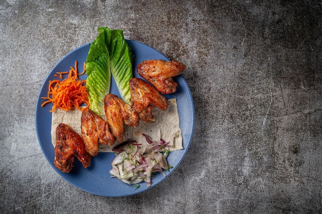 Подача блюда из меню ресторана. Куриные крылышки, наггетсы на гриле с тертой морковью и зеленью на тарелке на фоне серого каменного стола, вкуснейший шашлык