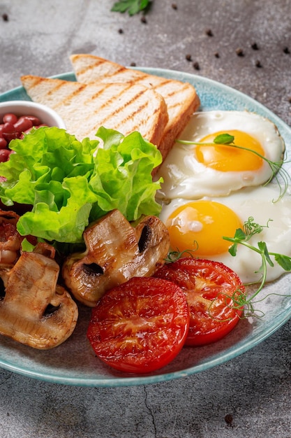 Serving a dish from the restaurant menu. Delicious and healthy breakfast with fried eggs, beans, sausages, grilled vegetables, tomatoes and mushrooms with salad on a plate against a gray stone table