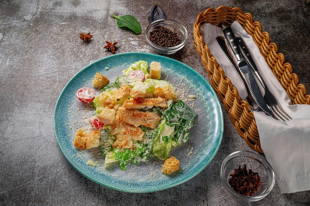 Подача блюда из меню ресторана. Свежий салат Цезарь из курицы с гренками, помидорами и зеленью на синей тарелке на фоне серого каменного стола, восхитительная закуска