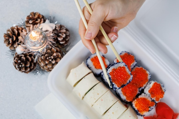 お祝いのスタイルで自宅のキッチンでプラスチックの箱にアジア ロールのセット女の子は箸で食べ物を保持していますクリスマス コンセプト新年の装飾