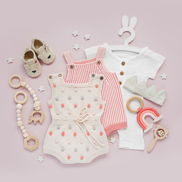 ピンクの背景にベビー服とアクセサリーのセット。さまざまなロンパース、ボディスーツのベビーシューズやおもちゃ。ファッション新生児。フラットレイ、上面図