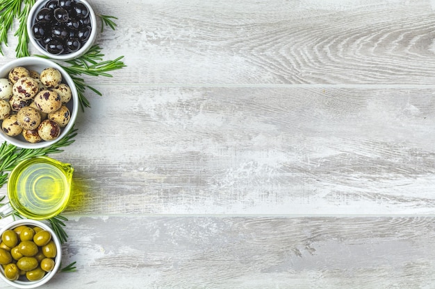 Set of black and green olives quail eggs on plates olive oil and rosemary on a light gray wooden background Top view copy space