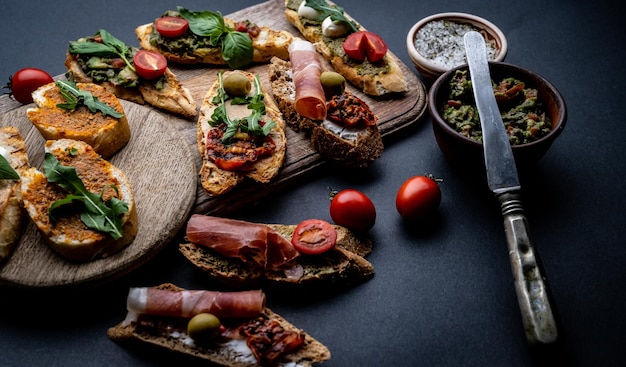 Set of bruschettas with jamon, olives, pesto, tomatoes, basil and mozzarella served on wooden board with guacamole and salt with herbs. Traditional mediterranean antipasti food composition