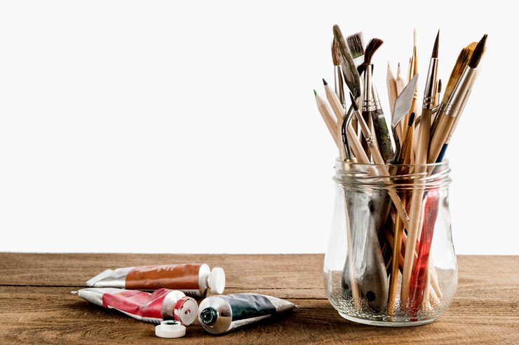 set of brushes in a jar next to oil paints on a wooden table on a white background