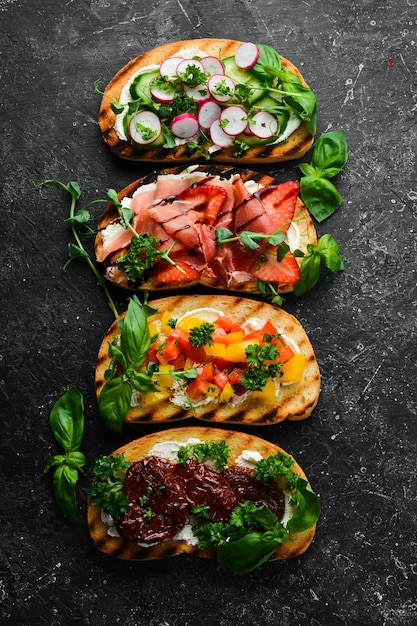 Set of colored sandwiches with tomatoes prosciutto vegetables and cheese on a black stone background Bruschetta Top view