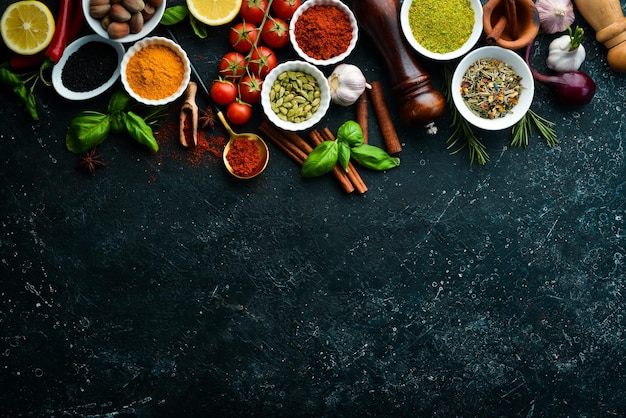 Set of colored spices in bowls and herbs on a black stone background. View from above. Top view.