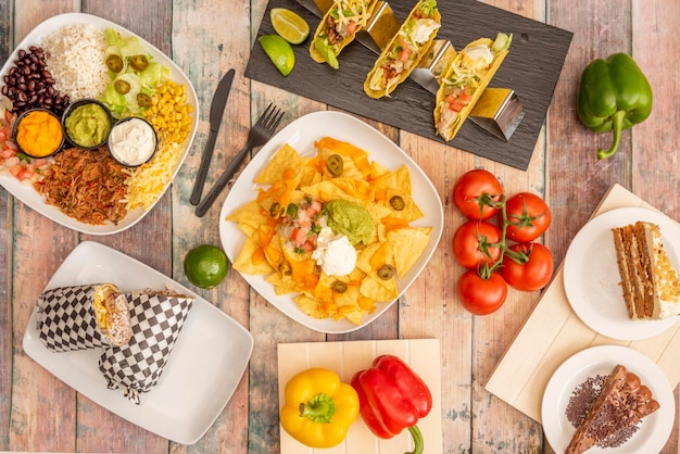 Set of colorful Mexican dishes with corn nachos avocado guacamole sweet corn cheddar cheese pico de gallo crispy corn tacos string tomatoes full burrito tricolor peppers on wooden table