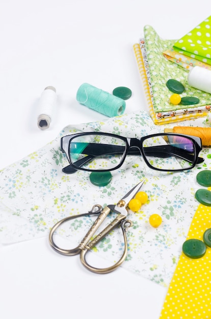 Set of colorful yellow and green fabrics, scissors, buttons, spools of thread and glasses