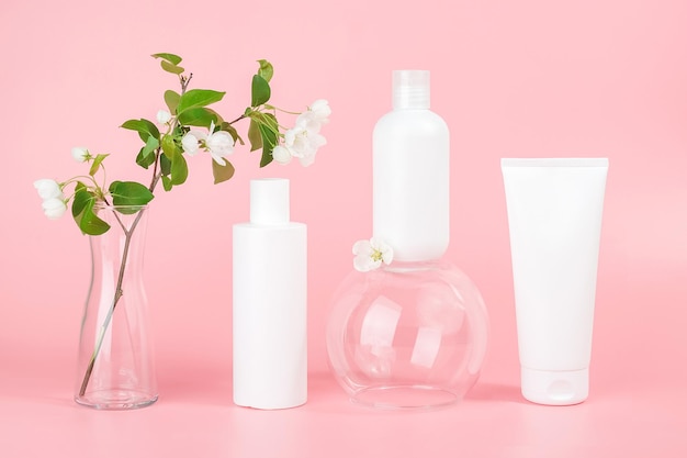 Set of cosmetic for skin care face body White blank cosmetics bottles and tube on glass podium and flowering branch in vase pink background Natural Organic Spa Cosmetic Beauty Concept Mockup