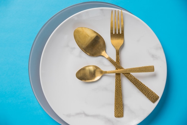 Set of cutlery spoon, fork, plate on blue wall. White marble plate. Top view .Table setting with gold silverware . styled elegant eating place setting