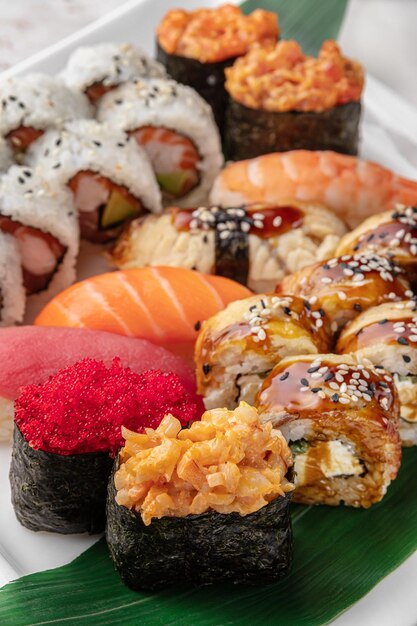 Set of different colorful maki sushi rolls nigiri and gunkan\
with tiger shrimp tomago salmon tuna chuka salad flying fish caviar\
tuna and green bamboo leaf in a white ceramic plate closeup