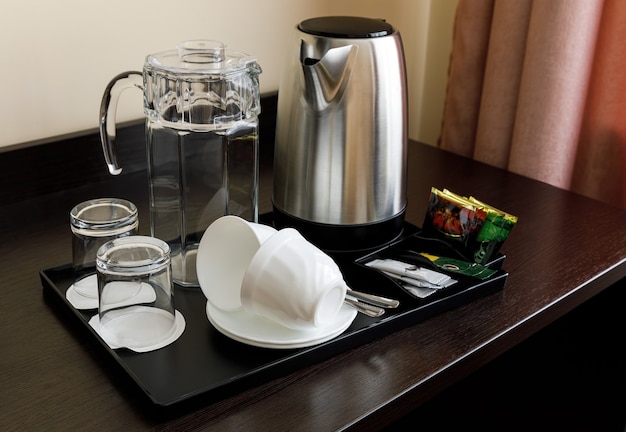 A set of dishes on a black tray for tea and coffee. Teapot, glass jug, glass glasses, cups. The table is dark wooden. Hotel, house.