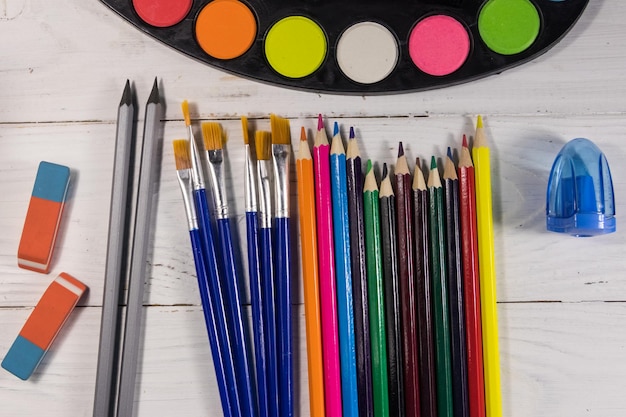 Set of drawing tools on white wooden background. Watercolor paints, paint brushes, color pencils, erasers and sharpener on desk. Top view