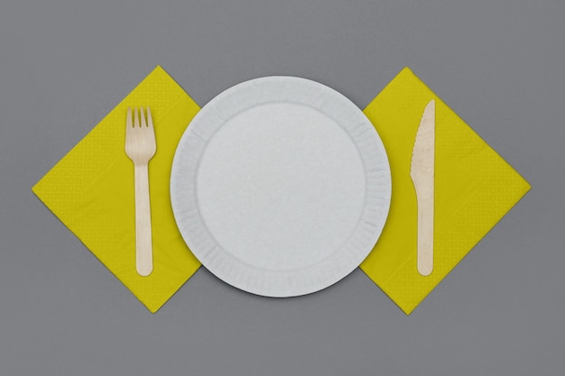 Set of eco friendly disposable tableware from natural material. white paper dish, wooden fork and knife and yellow napkins on gray background, top view. ecological concept