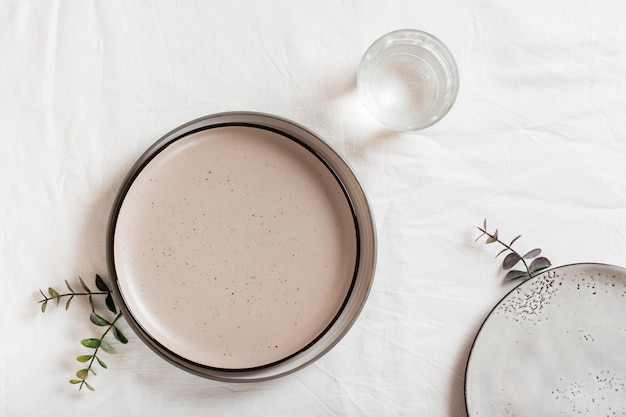 A set of empty ceramic plates a glass of water and eucalyptus branches on a white cloth Top view Ecofriendly concept