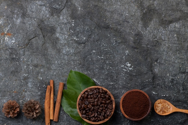 Set flat lay coffee beans in wood cup on green leaf, sugar in wood spoon, pine on black stone