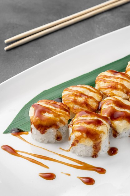Set of hot baked ricco rolls with shrimp mozzarella nori rice and green bamboo leaf in a white ceramic plate with chopstick on a dark gray textured background side view closeup