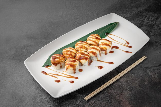 Set of hot baked ricco rolls with shrimp mozzarella nori rice and green bamboo leaf in a white ceramic plate with chopstick on a dark gray textured background side view