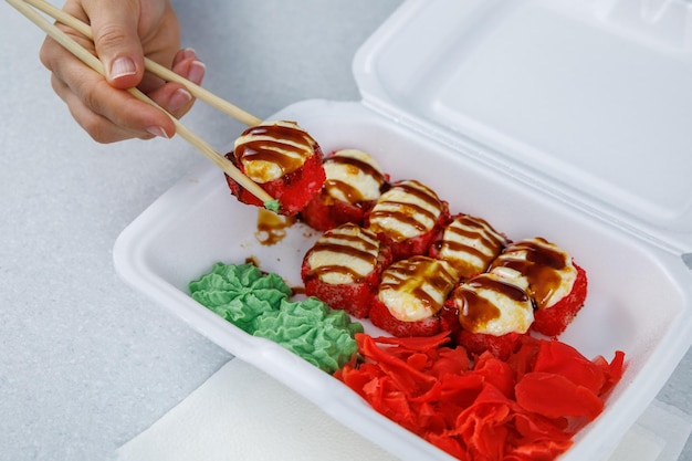 가벼운 테이블 위에 있는 플라스틱 상자에 일본 롤 세트 소녀는 젓가락으로 음식을 먹습니다 흰색 용기에 빠른 배달 스시